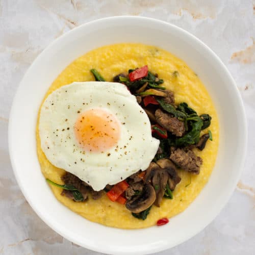 Cheesy polenta in a bowl topped with sausage sauteed veggies and a sunny side up egg