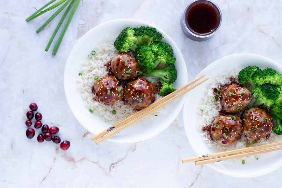 two bowls of cranberry chicken meatballs on a bed of spiced white rice with cranberry teriyaki sauce on top and broccoli on the side