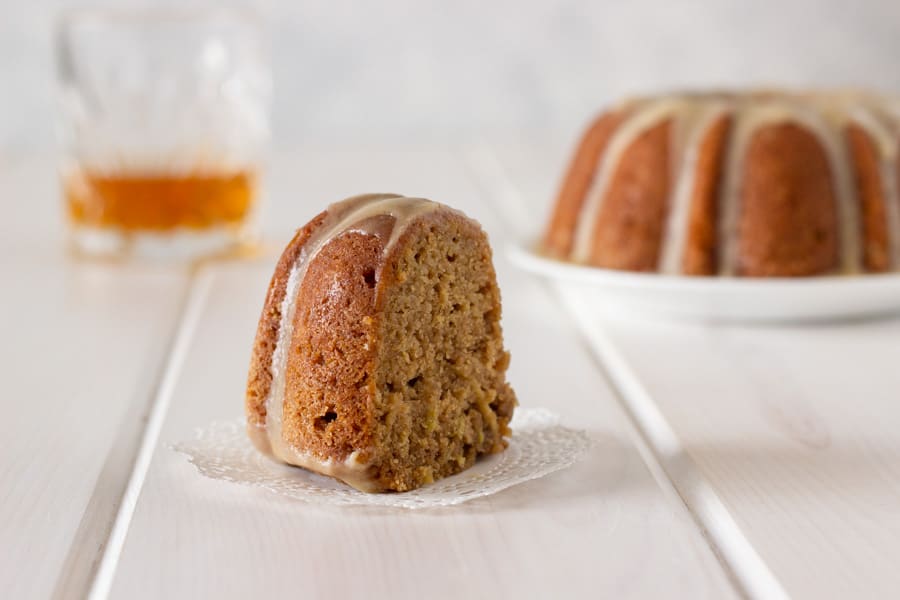 a piece of whole wheat apple Bundt cake with whiskey glaze with a whole cake and a glass of whiskey in the background