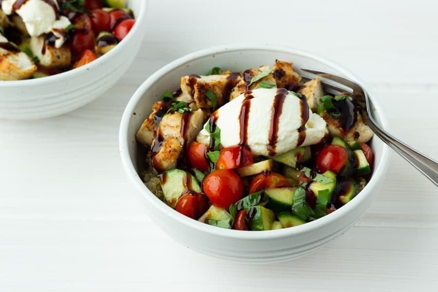 a white bowl with quinoa tomatoes, cucumbers, chicken, ricotta cheese basil and balsamic drizzle