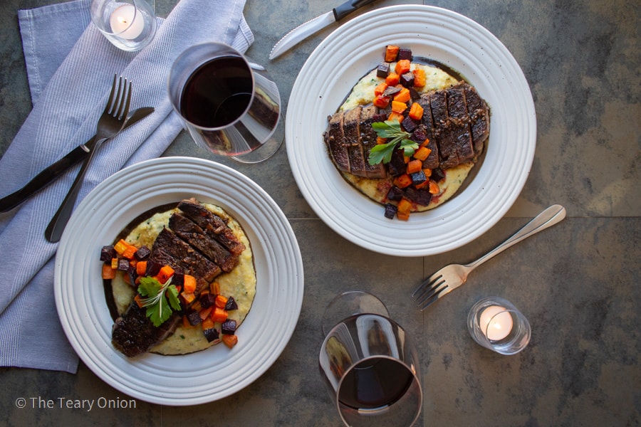 two plates of steak and polenta with roasted root vegetables on a table with two glasses of wine and two candles