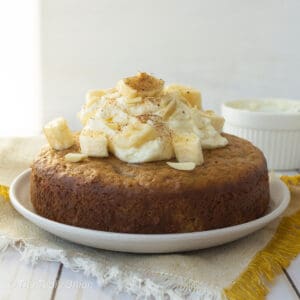 A round banana cake on a white plate with whipped cream and bananas on top.